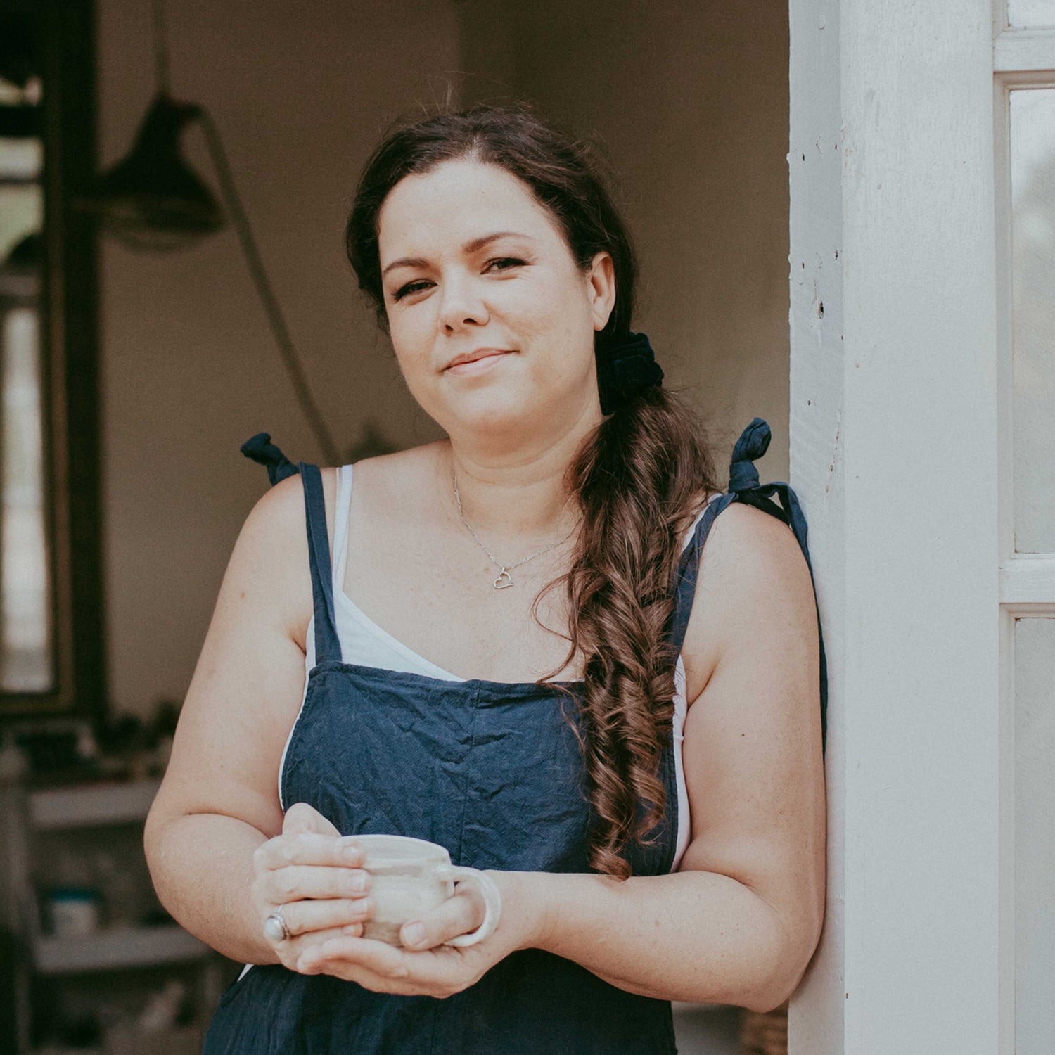 A photo of Hayley owner of Earth and Nectar standing in the doorway of her pottery studio holding a handmade stoneware mug she has made
