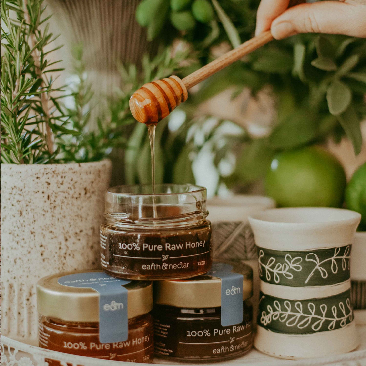 Three jars of Earth and Nectar honey sitting on top of a tray surrounded by Earth and Nectar Property. One of the honey jars is open a honey dipper is being held showing the pure, raw Western Australian honey drizzle from the dipper into the jar